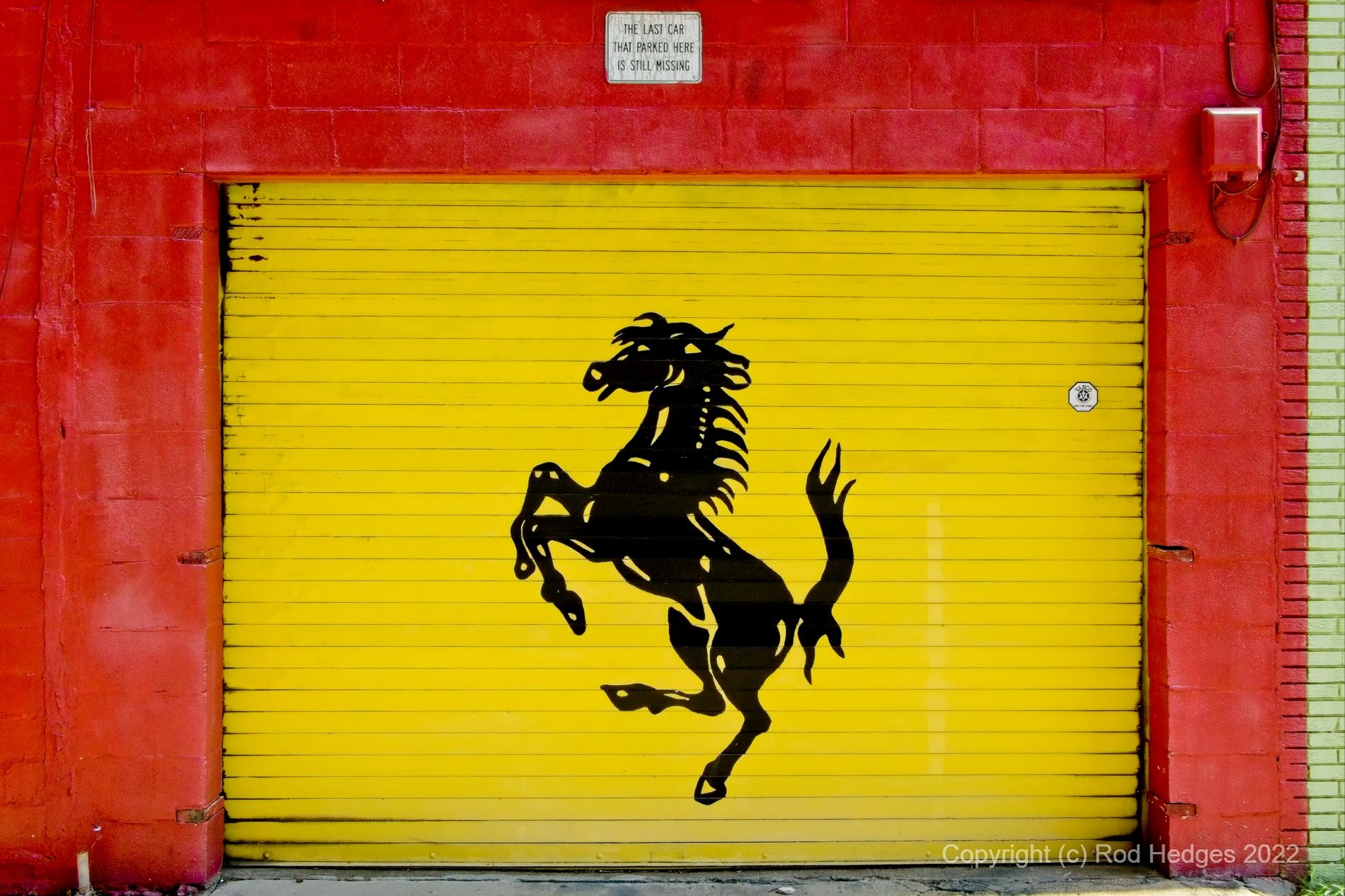 This mural features a silhouetted maverick horse in fighting pose centered upon a bright yellow wooden garage door. A small "no parking" sign hangs above the door and displays the meme "The last car that parked here is still missing." The mural is framed by the building's wall of bright red cinderblocks.